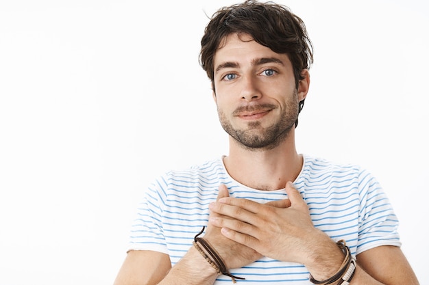 Disparo de cintura para arriba de un joven apuesto joven feliz y conmovido con ojos azules y cerdas tomados de la mano en el pecho sonriendo y mirando agradecido con aprecio al frente sobre la pared gris