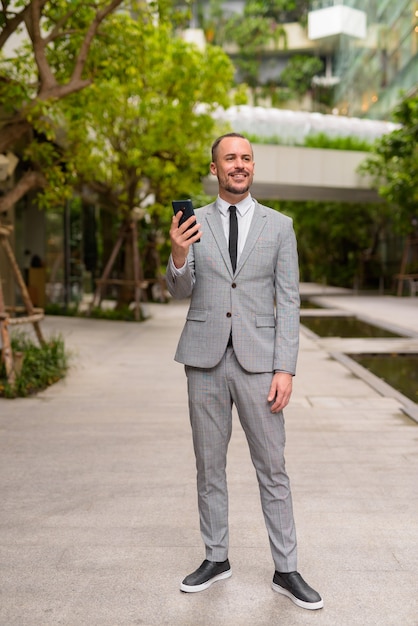 Foto disparo de cuerpo completo del empresario barbudo calvo hispano feliz pensando y usando el teléfono con la naturaleza en la ciudad