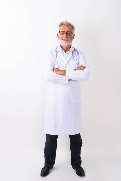 Disparo de cuerpo completo de feliz senior hombre barbudo doctor sonriendo mientras está de pie con los brazos cruzados sobre blanco