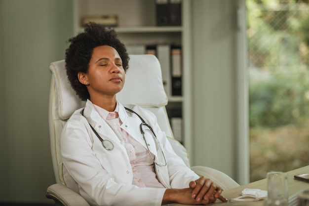 Disparo de una doctora africana cansada sentada sola con los ojos cerrados en el escritorio de su sala de consulta durante la pandemia de COVID-19.