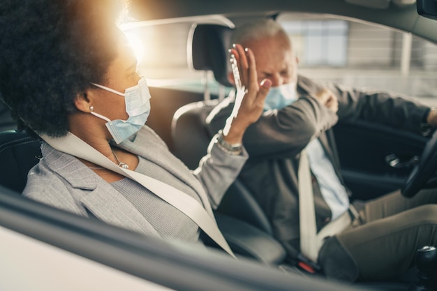 Disparo de dos exitosos empresarios multiétnicos con máscara protectora que viajan en automóvil durante la pandemia de COVID-19.