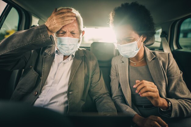 Disparo de dos personas multiétnicas preocupadas con máscara protectora usando una computadora portátil y discutiendo mientras estaban sentados en el asiento trasero de un automóvil en su viaje de negocios durante la pandemia de COVID-19.