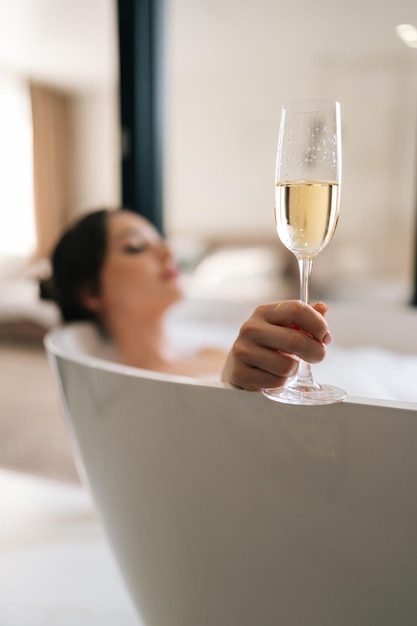 Disparo de enfoque selectivo vertical de una joven mujer relajada tendida en un baño de espuma sosteniendo champán de vidrio disfrutando de una bebida alcohólica durante una recreación lujosa para el fin de semana