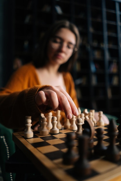Disparo de enfoque selectivo vertical de una mujer joven con anteojos elegantes haciendo que el ajedrez se mueva sentado en un sillón en una habitación oscura