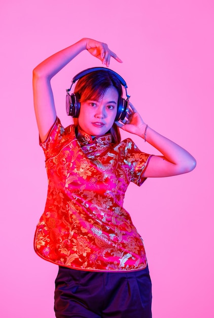 Disparo de estudio de modelo de mujer asiática milenaria en rojo chino tradicional cheongsam camisa qipao ojos cerrados de pie escuchando música a través de auriculares bailando manos levantadas sobre fondo rosa.