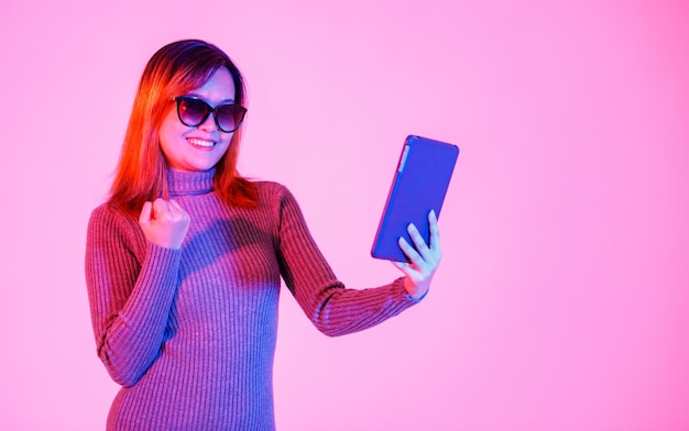 Foto disparo de estudio de modelo de mujer joven asiática en suéter de cuello alto gris en tonos negros de pie sonriendo sosteniendo tablet pc sobre fondo rosa claro.