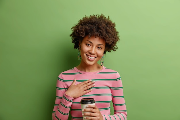 Disparo de estudio de mujer agradecida y complacida mantiene la mano en el pecho escucha palabras conmovedoras sostiene una taza de café desechable inclina la cabeza usa un jersey de rayas aislado sobre una pared verde vivo