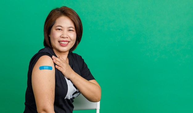 Foto disparo de estudio de una paciente asiática de mediana edad que se sienta, sonríe y muestra un vendaje de yeso azul en el brazo después de recibir la vacunación contra el coronavirus covid 19 por parte del médico en la clínica sobre fondo verde.