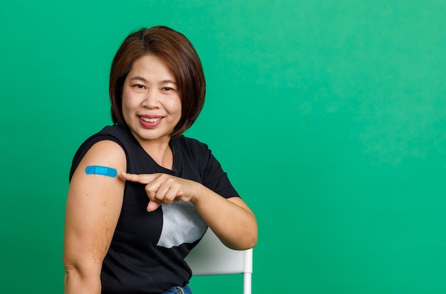 Disparo de estudio de una paciente asiática de mediana edad que se sienta, sonríe y muestra un vendaje de yeso azul en el brazo después de recibir la vacunación contra el coronavirus covid 19 por parte del médico en la clínica sobre fondo verde.