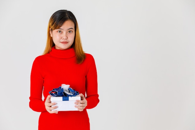 Disparo de estudio de retrato de feliz cumpleañera asiática joven en vestido de suéter rojo de manga larga con caja de regalo con cinta azul plateada en las manos celebrando el día importante sobre fondo blanco.