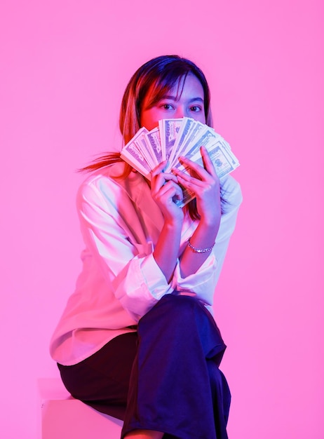 Disparo de estudio de retrato de modelo de mujer de negocios asiática rica riqueza en casual viste sentado mirar a la cámara sosteniendo la pila de moneda de billetes en efectivo de cien dólares USD sobre fondo rosa claro.