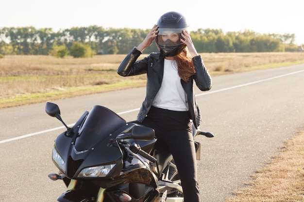 El disparo exterior de una motociclista rápida femenina usa casco protector, posa en moto, se para en moto, recorre largas distancias, tiene un viaje inolvidable. Personas, equitación, seguridad y concepto extremo.