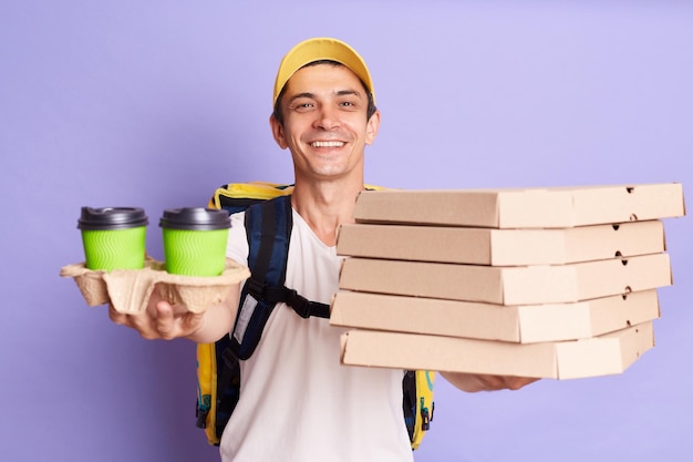 Disparo horizontal de un apuesto repartidor con gorra amarilla y camiseta sosteniendo pizza en cartón y café para llevar complacido con el cliente aislado de fondo violeta