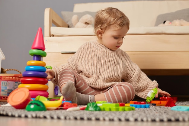 Disparo horizontal de un bebé con suéter elegante jugando con coloridos juguetes de arco iris sentados en el suelo con juguetes educativos de mirada atenta para el desarrollo temprano de los niños