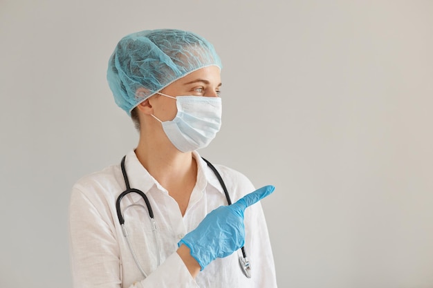 Disparo horizontal de doctora con gorro médico, guantes, mascarilla quirúrgica y bata, mostrando una maqueta de texto promocional o su anuncio con el dedo índice.