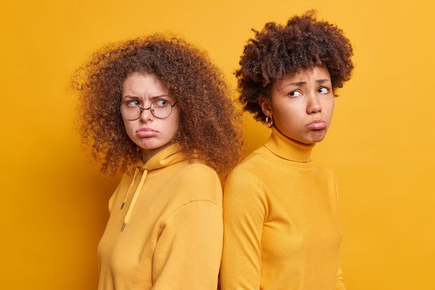 Disparo horizontal de dos infelices mujeres diversas que están una al lado del otro y no hablan después de la pelea, los labios del bolso vestidos con ropa casual aislada sobre una pared amarilla. Concepto de emociones negativas