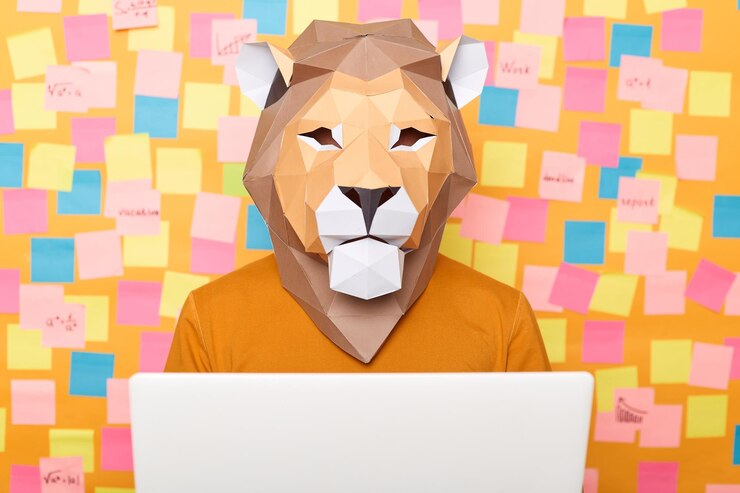 Foto disparo horizontal de un hombre anónimo con un jersey naranja y una máscara de león de papel posando contra una pared amarilla con pegatinas trabajando en una laptop sentada frente a la pantalla del portátil