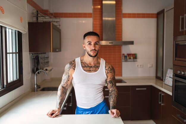 Disparo horizontal de un joven guapo tatuado posando en la cocina