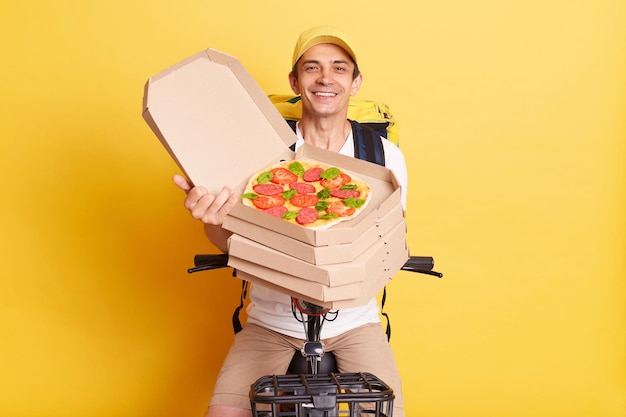 Disparo horizontal de un mensajero alegre que sostiene una caja de pizza de cartón abierta que muestra el pedido al cliente con una camiseta blanca y una gorra aisladas sobre fondo amarillo