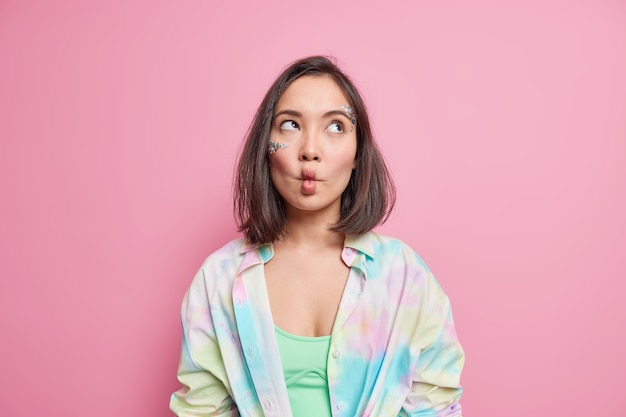 Disparo horizontal de mujer asiática pensativa hace tontos labios de pez alrededor concentrado arriba vestido con camisa colorida aislada sobre pared rosa siendo infantil. Concepto de expresiones faciales