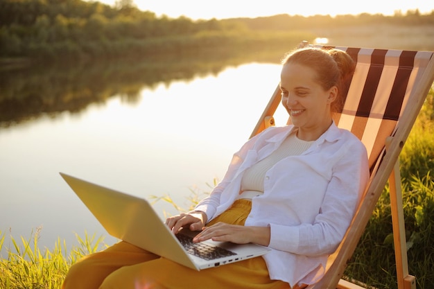 Disparo horizontal de una mujer caucásica sonriente con camisa blanca sentada junto al agua en una silla plegable y trabajando en una computadora portátil escribiendo en un portátil trabajos independientes durante sus vacaciones con una hermosa vista