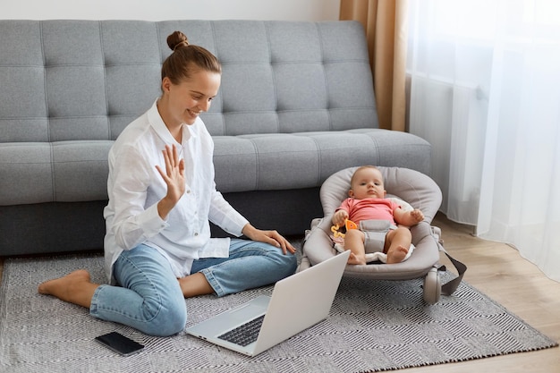Disparo horizontal de una mujer encantada con peinado de moño con camisa blanca y jeans sentados en el suelo con un bebé recién nacido y haciendo una videollamada en una laptop saludando con la mano
