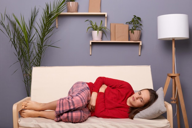 Disparo horizontal de una mujer enferma triste y malsana que usa suéter rojo y pantalones a cuadros acostados en un sofá en el interior de su casa que sufre dolor de estómago sosteniendo su vientre siente dolor menstrual