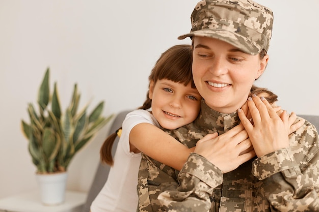 Disparo horizontal de una mujer extremadamente feliz y satisfecha con uniforme de camuflaje y gorra posando con su hija abrazando a su madre militar que llegó a casa de la guerra o del ejército