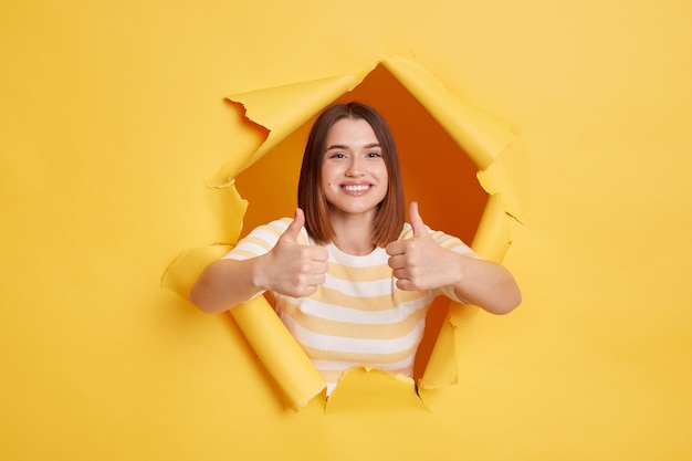 Disparo horizontal de mujer feliz sonriente con camiseta a rayas mirando a través de papel amarillo rasgado mostrando el pulgar hacia arriba gesto de aprobación buena retroalimentación