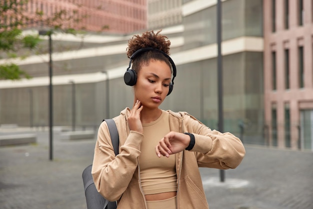 Disparo horizontal de una mujer seria de cabello rizado que revisa el pulso después de que el entrenamiento cardiovascular mira la pantalla de smartwacth vestida con ropa deportiva escucha música a través de auriculares inalámbricos que usa puestos de karemat al aire libre