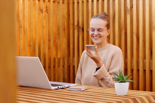 Disparo horizontal de una mujer sonriente y encantadora con un peinado de moño sosteniendo un teléfono inteligente y grabando un mensaje de voz usando un suéter beige trabajando en una computadora portátil contra una pared de madera