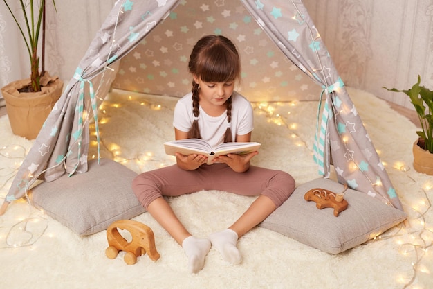 Disparo horizontal de una niña con una camiseta blanca leyendo un libro con placer y mirando fotos con interés sentada en el juguete infantil wigwam el niño está en casa