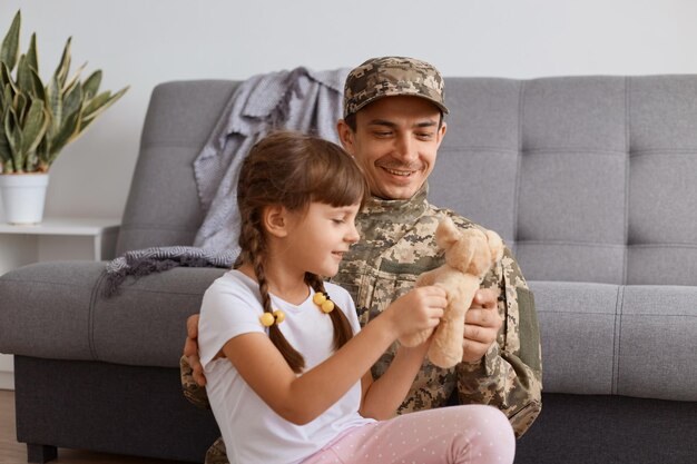 Disparo horizontal de padre encantado con su hijo, hombre soldado caucásico con uniforme de camuflaje que regresa a casa después del ejército, disfrutando del tiempo con su pequeña hija, el niño mostrando su juguete a papá.