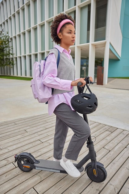 Disparo de longitud completa de una adolescente de pelo rizado vestida con ropa de calle lleva una mochila paseos en scooter eléctrico en la calle durante el tiempo libre durante el fin de semana