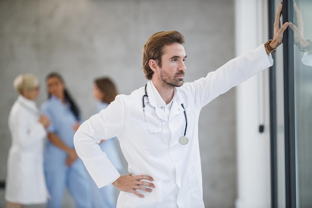 Disparo de un médico cansado de pie y pensativo mirando por la ventana mientras tomaba un descanso rápido en un pasillo del hospital durante la pandemia de Covid-19.