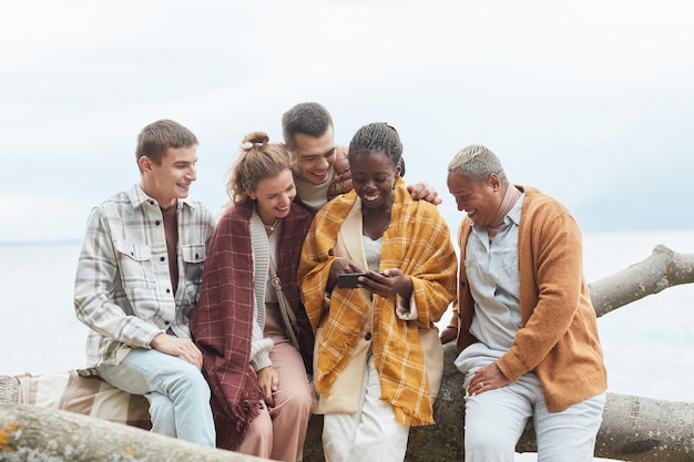 Disparo mínimo de un grupo diverso de jóvenes en la playa en otoño juntos usando el espacio de la copia del teléfono inteligente