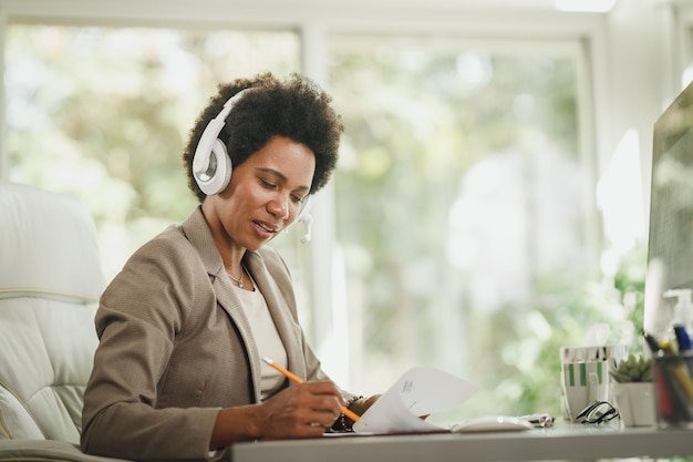 Disparo de una mujer africana que usa auriculares y trabaja en su escritorio durante la pandemia del virus de la corona.