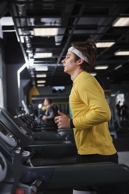 Disparo de perfil vertical de un atleta masculino con diadema corriendo en la cinta de correr en el gimnasio