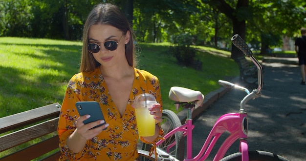 Disparo en primer plano. Retrato de mujer joven atractiva con una bicicleta utiliza smartphone y bebiendo limonada en un banco del parque. La mujer joven de moda tiene estilo de vida activo y deportes.