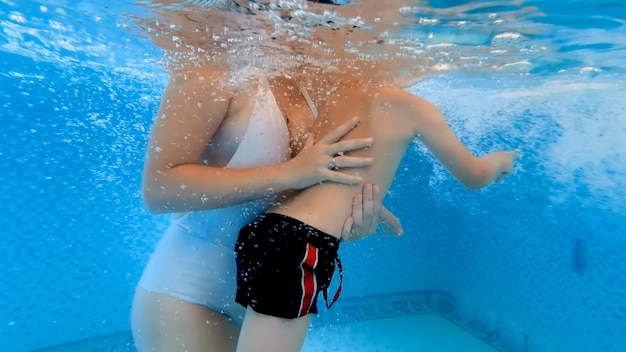 Disparo subacuático joven madre apoyando a su pequeño hijo aprendiendo a nadar en la piscina. Concepto de salud familiar y deportiva.