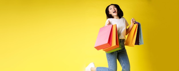Disparo de tamaño completo de hermosa mujer asiática saltando con bolsas de compras y sonriendo feliz backgrou amarillo