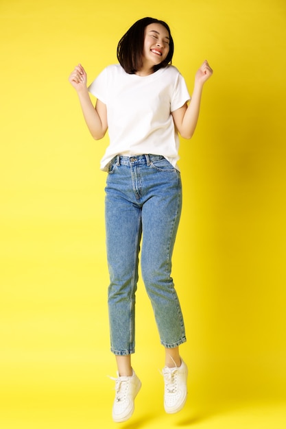 Disparo de tamaño completo de mujer asiática sin preocupaciones saltando y bailando, divirtiéndose, posando en jeans y camiseta blanca sobre fondo amarillo.