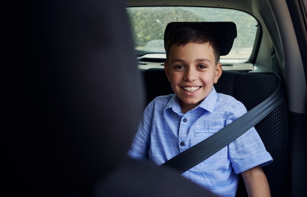 Foto disparo a través del asiento del conductor de un niño de escuela sonriente abrochado con el cinturón de seguridad mientras viaja en automóvil en el asiento de seguridad para niños