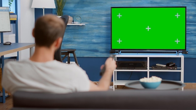 Disparo de trípode estático del hombre cambiando de canal mientras mira la pantalla verde en la televisión y se sienta en el sofá. Vista sobre el hombro de la persona que se relaja usando el zapping remoto de la televisión en la pantalla de croma.