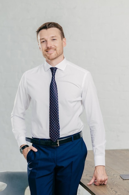 Foto disparo vertical de un apuesto hombre alegre sin afeitar que usa ropa formal mantiene la mano en el bolsillo cerca de la mesa de la oficina tiene una sonrisa positiva hombre de negocios próspero en el lugar de trabajo concepto de negocio