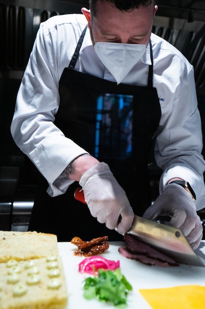 Foto disparo vertical de un chef masculino con una máscara facial cortando una carne a la parrilla