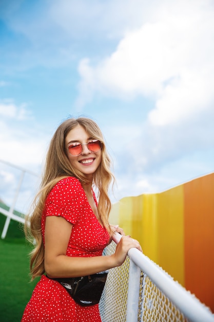 Disparo vertical de una hermosa chica rubia con vestido de verano y gafas de sol rojas, apoyado en la barandilla en el parque.