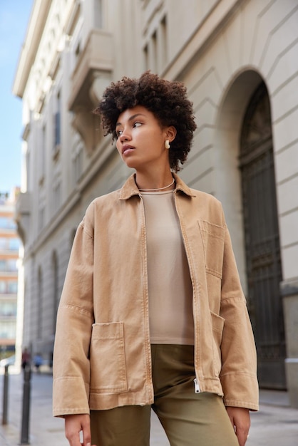 Disparo vertical de una hermosa mujer rizada pensativa vestida con ropa de calle camina en la ciudad antigua concentrada en poses contra los paseos de edificios urbanos afuera durante el día. Concepto de estilo de vida.