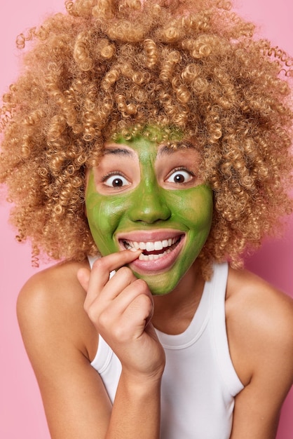 Disparo vertical de una joven rubia feliz y rizada que aplica una máscara de pepino verde, sonríe ampliamente, muerde el dedo, usa una camiseta informal, posa en interiores Tratamientos faciales de belleza y concepto de procedimientos cosméticos