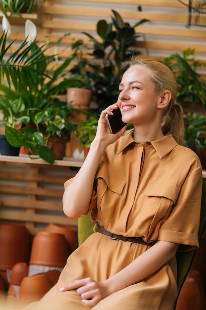 Disparo vertical de una joven sonriente propietaria de una tienda de flores sentada en una tienda de flores y hablando por teléfono inteligente con el cliente
